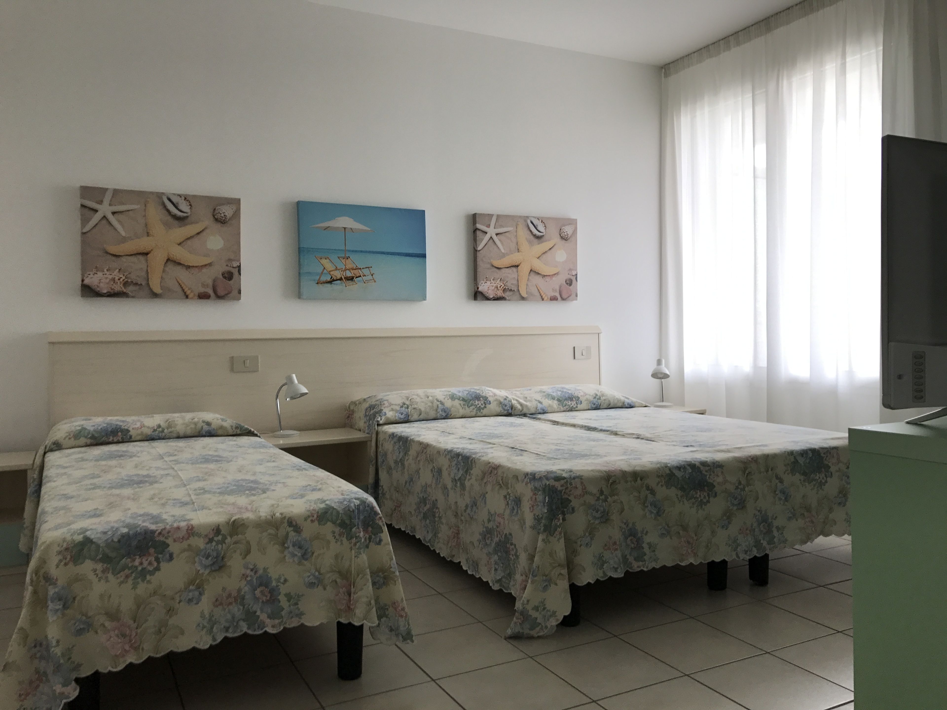 A hotel room with two beds, a desk, a chair, and a TV. There are three paintings on the wall, and a window with curtains.