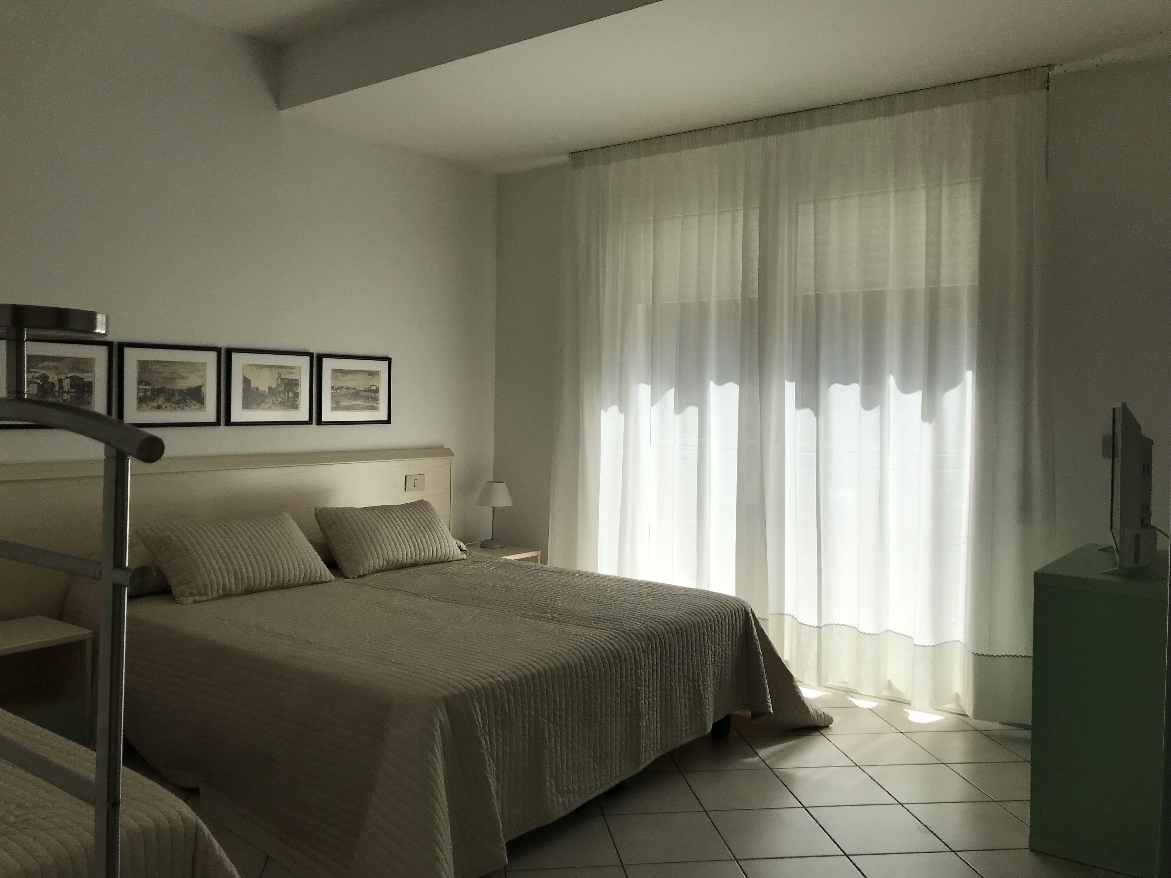 A hotel room with a large bed, a bedside table, a lamp, and a TV on a stand. There are three framed pictures on the wall above the bed.