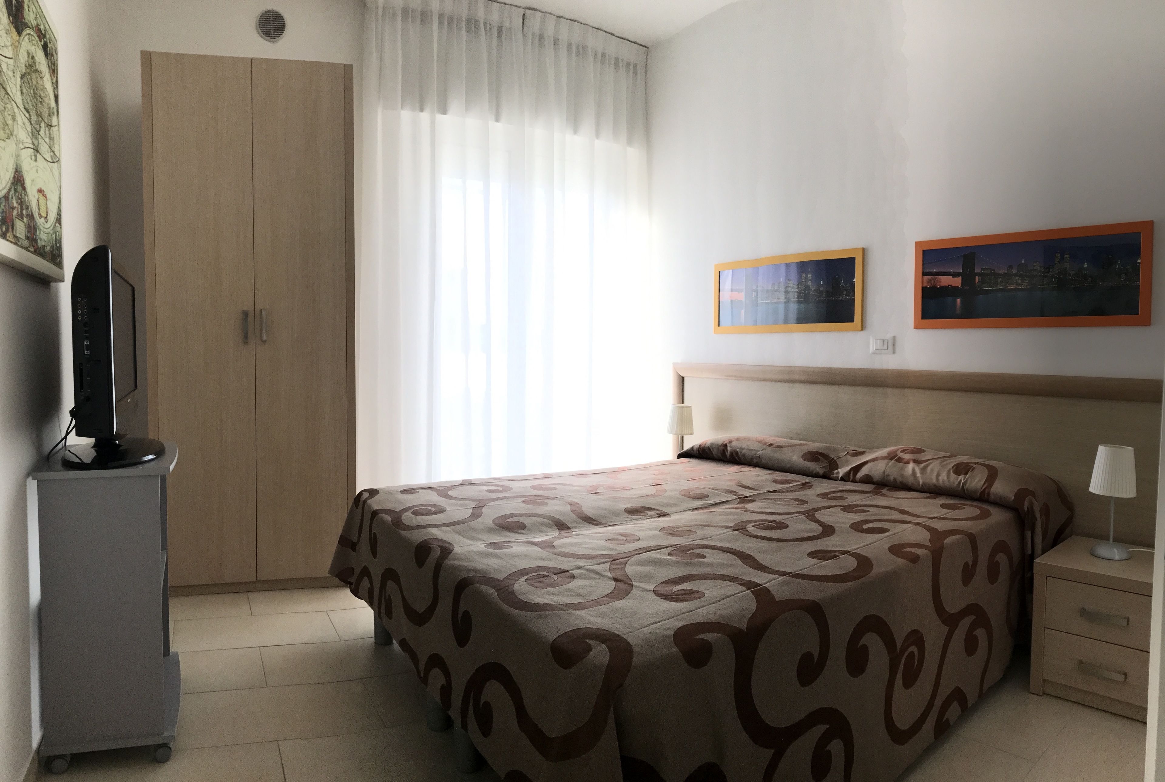 A hotel room with a bed, a desk, a chair, a wardrobe, and two framed pictures on the wall.