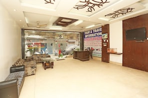 A hotel lobby with sofas, a reception desk, a ceiling fan, and a signboard with tour information.