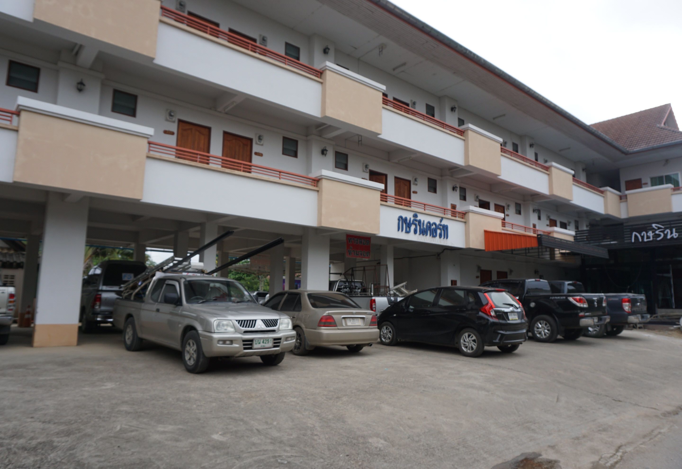 A multi-story building with a parking area. There are several cars parked in front, including a pickup truck and a sedan. The building has multiple balconies and a sign with Thai script.