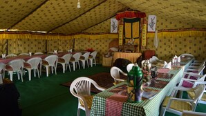 Un área de comedor con carpas, mesas y sillas, una mesa central con un termo verde y un tapiz decorativo.