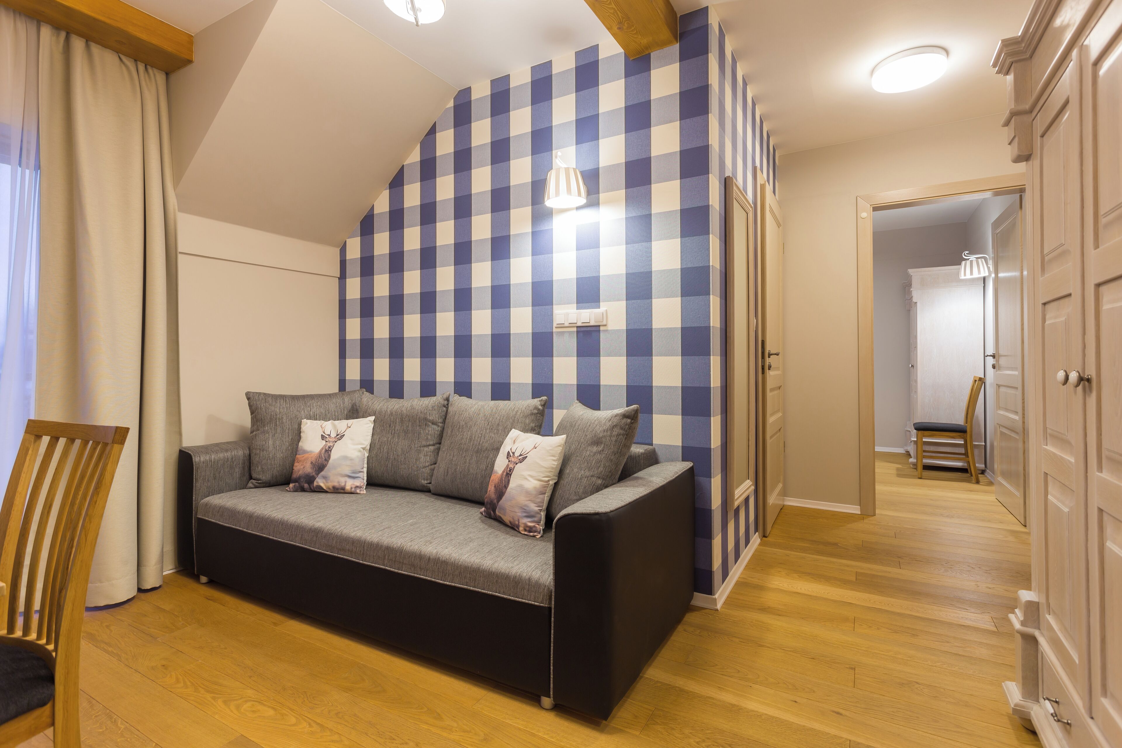 Una habitación con pared azul y blanca a cuadros, un sofá gris con cojines, una silla de madera y suelo de madera.