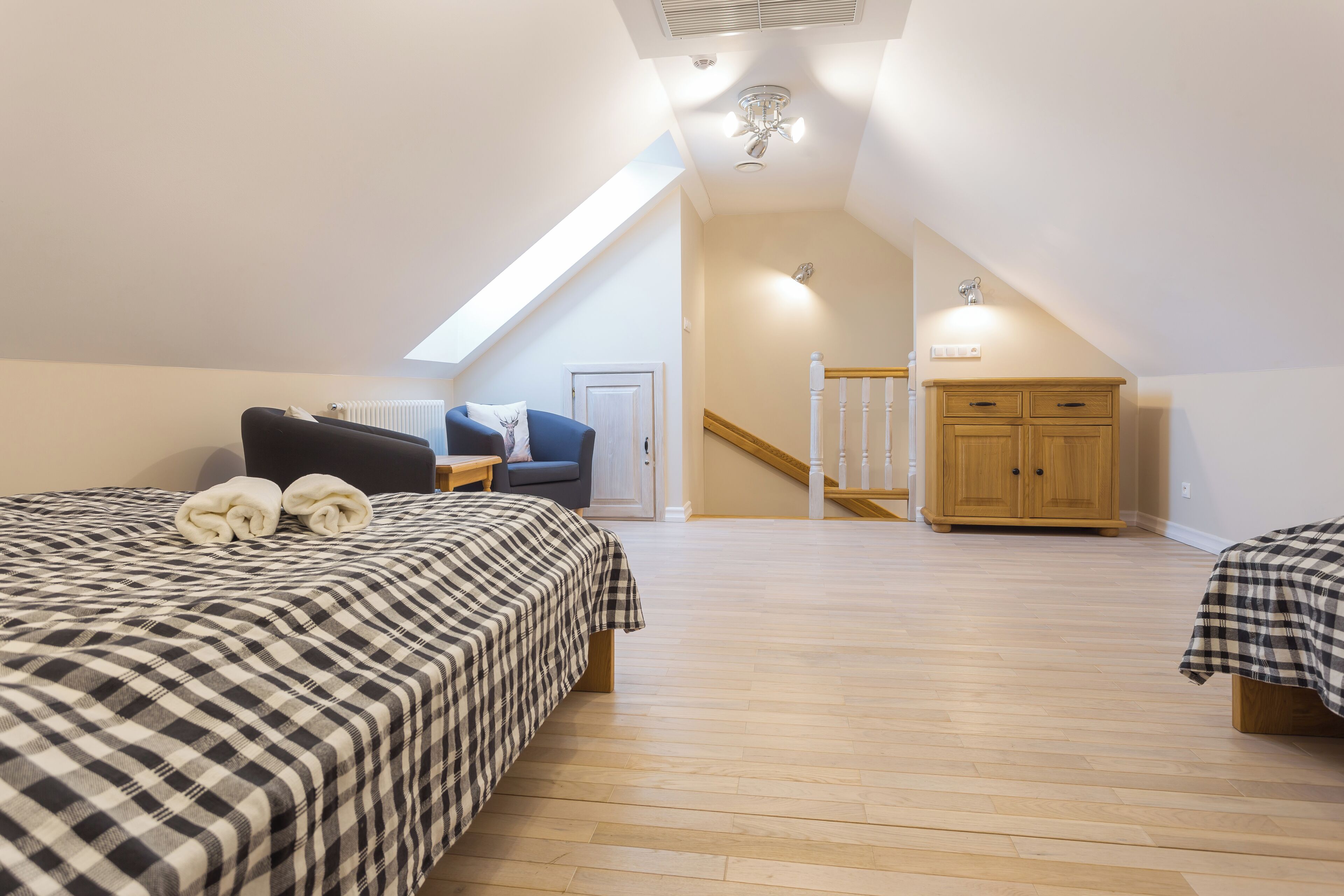 A bedroom with a sloped ceiling, a bed with a checkered bedspread, a blue chair, a wooden wardrobe, and a staircase leading to an upper floor.