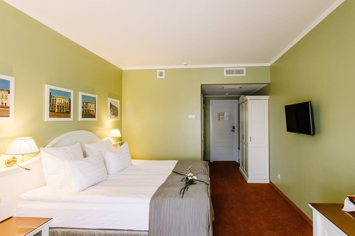 Hotellihuone, jossa on sänky, yöpöytävalaisimet, taulutelevisio ja seinällä olevia kehystettyjä tauluja.