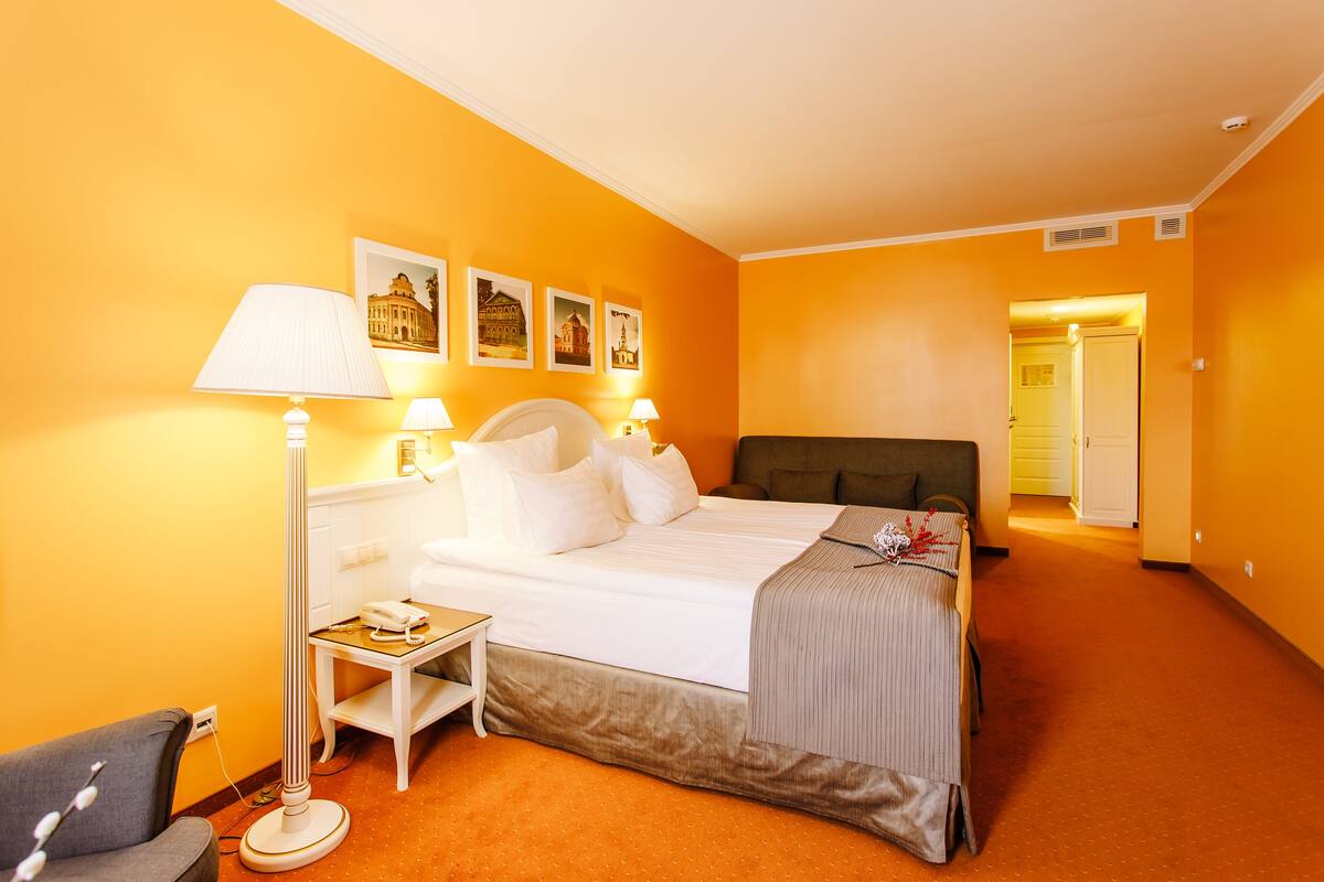 Hotellihuone, jossa on sänky, yöpöytälamput, yöpöytä, tuoli ja ovi, joka johtaa toiseen huoneeseen.
