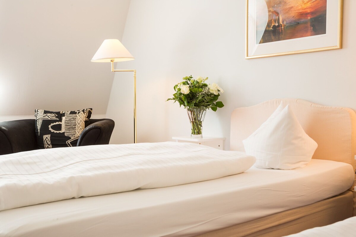 Ein ordentlich bezogenes Bett mit weißen Leinen, ein schwarzer Lederstuhl mit gemusterten Kissen, eine Stehlampe, eine Vase mit Blumen und ein gerahmtes Bild an der Wand.