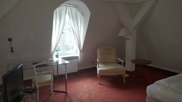 A room with a single bed, a chair, a desk with a computer, a lamp, and a window with curtains.