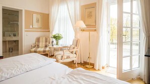Een slaapkamer met een groot bed, twee stoelen, een nachtkastje met een lamp, een spiegel en een raam met gordijnen.