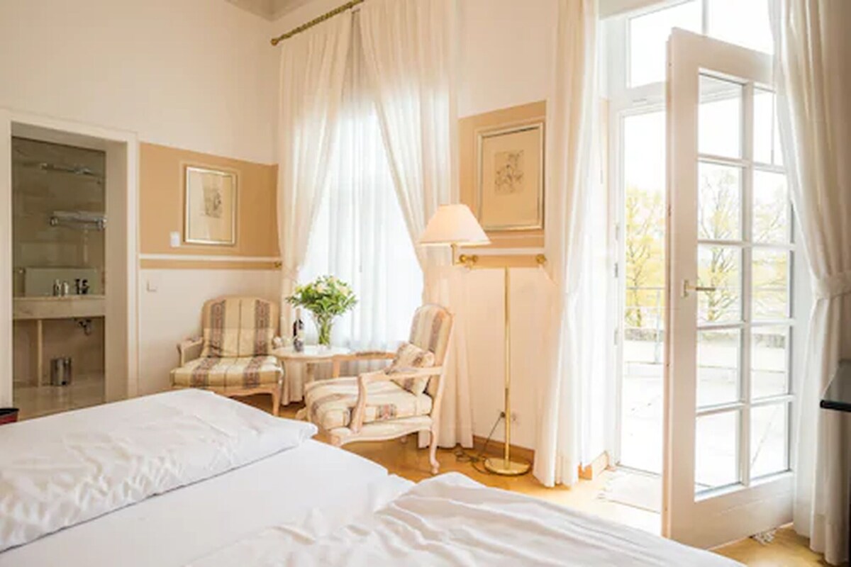Ein Schlafzimmer mit einem großen Bett, zwei Stühlen, einem Nachttisch mit Lampe, einem Spiegel und einem Fenster mit Vorhängen.