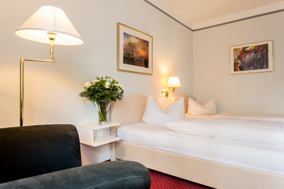 Ein Hotelzimmer mit einem Bett, zwei Wandlampen, einer Vase mit Blumen und zwei gerahmten Bildern an der Wand.
