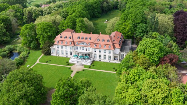 Een groot, historisch gebouw met een symmetrische gevel, omgeven door weelderig groen en een verzorgd gazon.