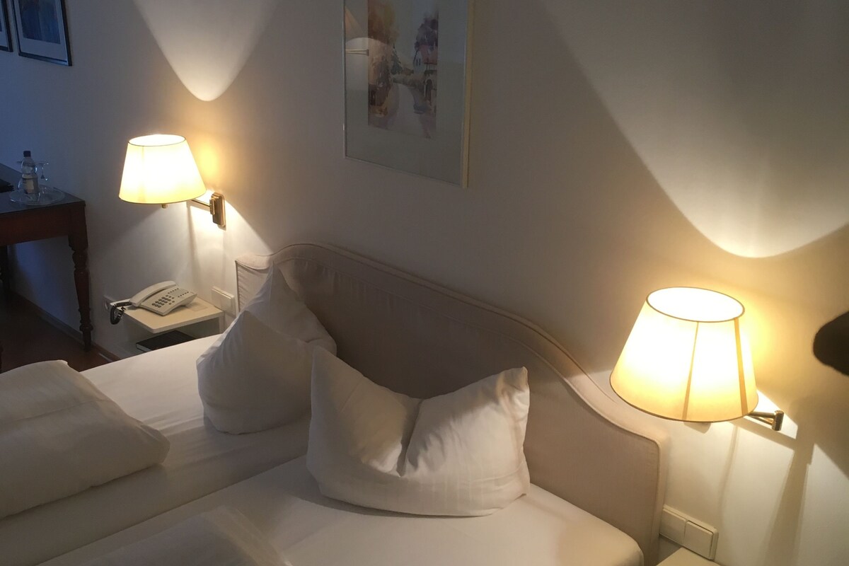Ein Hotelzimmer mit einem ordentlich bezogenen Bett, zwei Nachttischlampen, einem Telefon am Nachttisch und einem gerahmten Bild an der Wand.