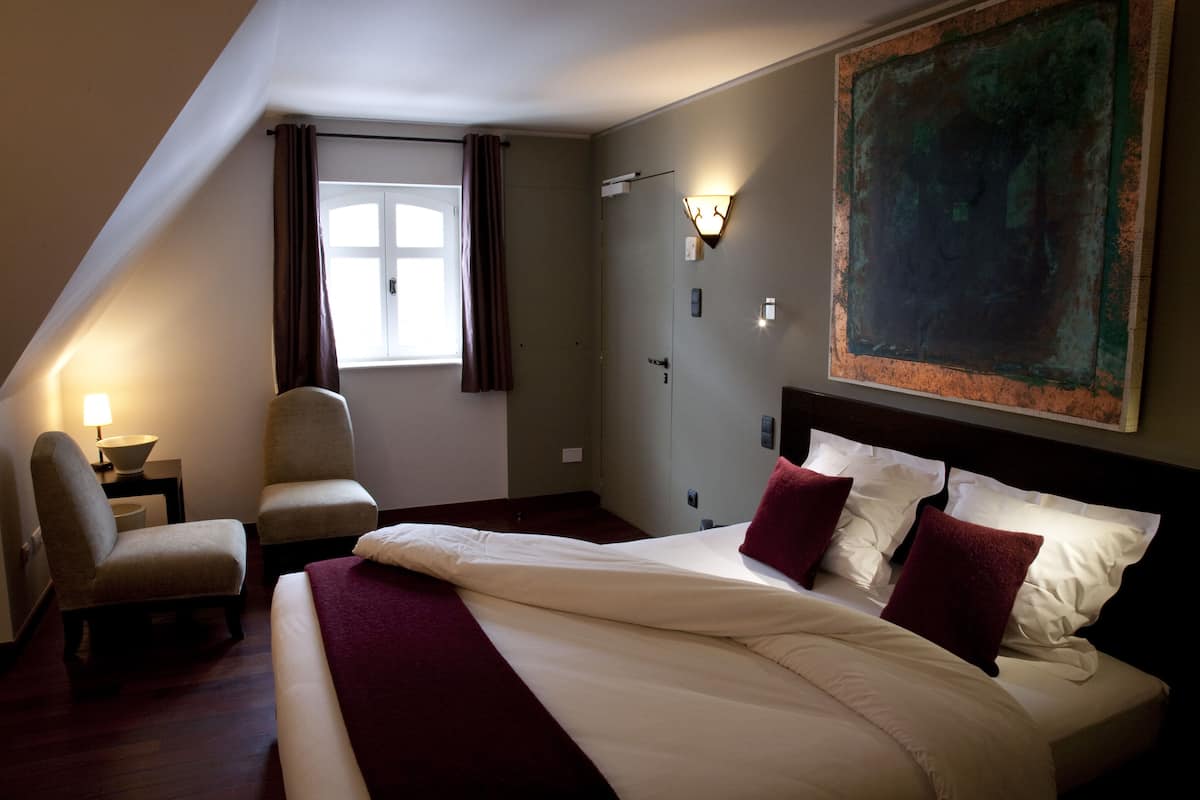 Een hotelkamer met een groot bed, een stoel, een nachtkastje, een raam met gordijnen en een schilderij aan de muur.