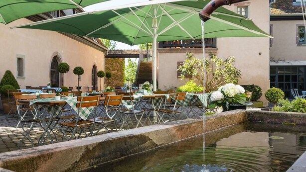 Un área de comedor al aire libre con mesas y sillas, cubierta por sombrillas verdes, y una pequeña fuente en primer plano.