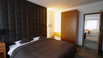 A modern hotel room with a large bed, a wooden wardrobe, a bedside table with a lamp, and a view into the adjacent room.