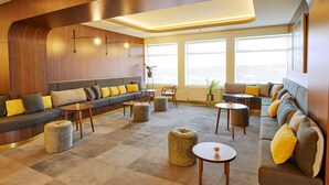 A modern lounge area with a curved wooden wall, a grey sofa with yellow cushions, round ottomans, and a small table with a candle. - Fosshotel Stykkisholmur (Stykkisholmur)