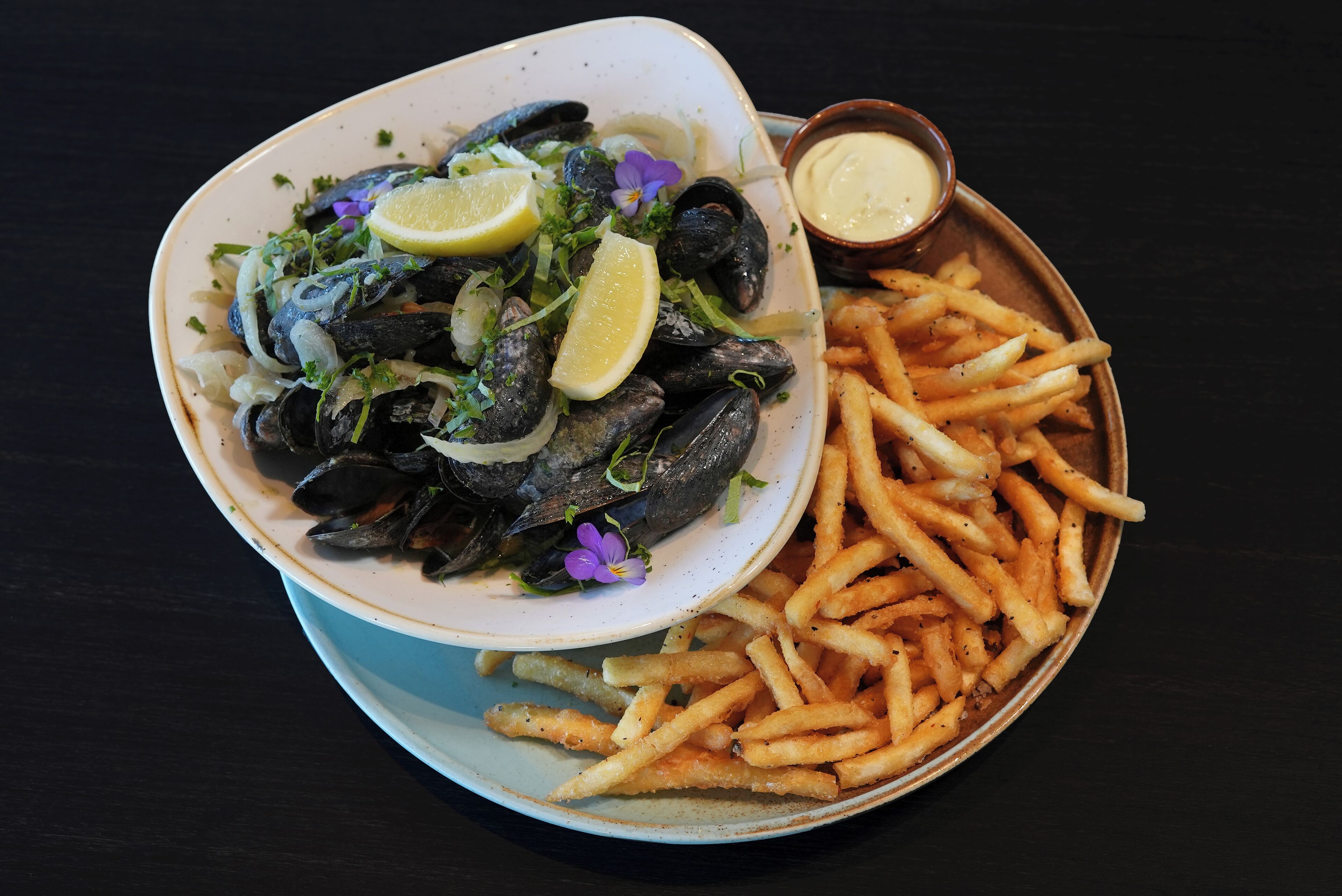 Un bol de mejillones con limón y hierbas, acompañado de patatas fritas sazonadas y un recipiente pequeño con salsa.