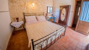 A bedroom with a stone wall, a bed with a quilt, a wooden door, and a small table with a lamp. - A Lumìa (Noto)