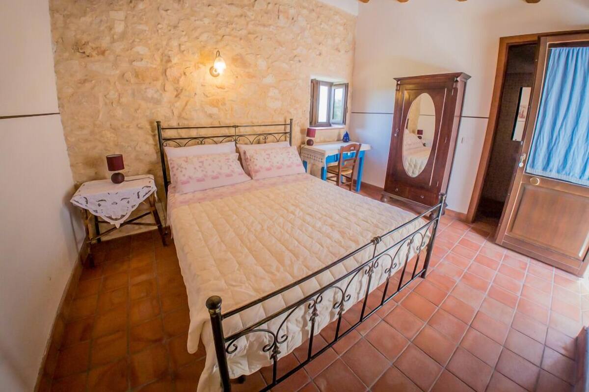Un dormitorio con pared de piedra, una cama con colcha, una puerta de madera y una mesita de noche con lámpara.