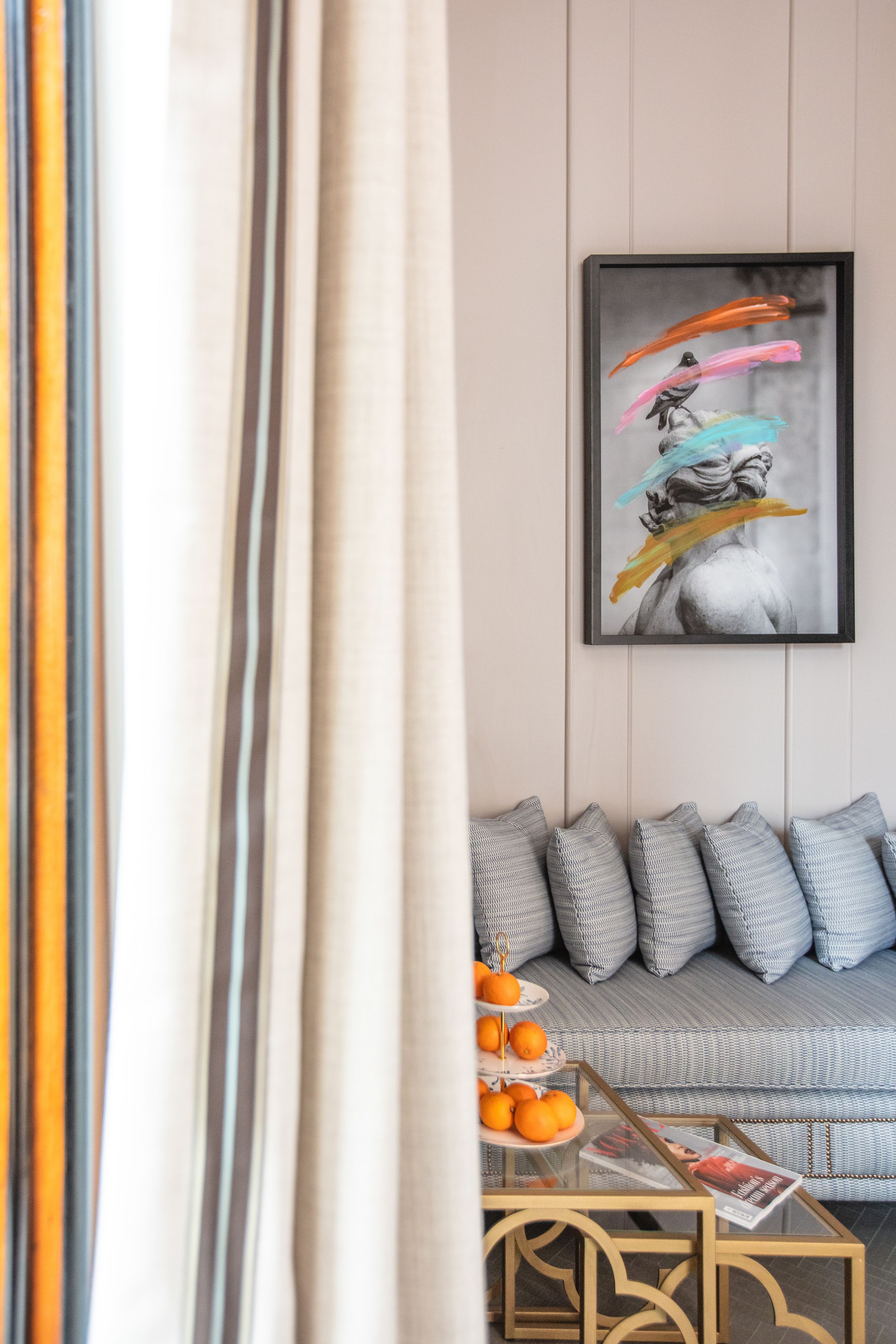 A living room with a sofa, cushions, a coffee table with oranges, and a framed artwork on the wall.