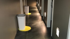 Un pasillo con suelo de baldosas oscuras, paredes blancas y puertas a ambos lados. Se observan dos botes de basura con base amarilla a lo largo del pasillo.