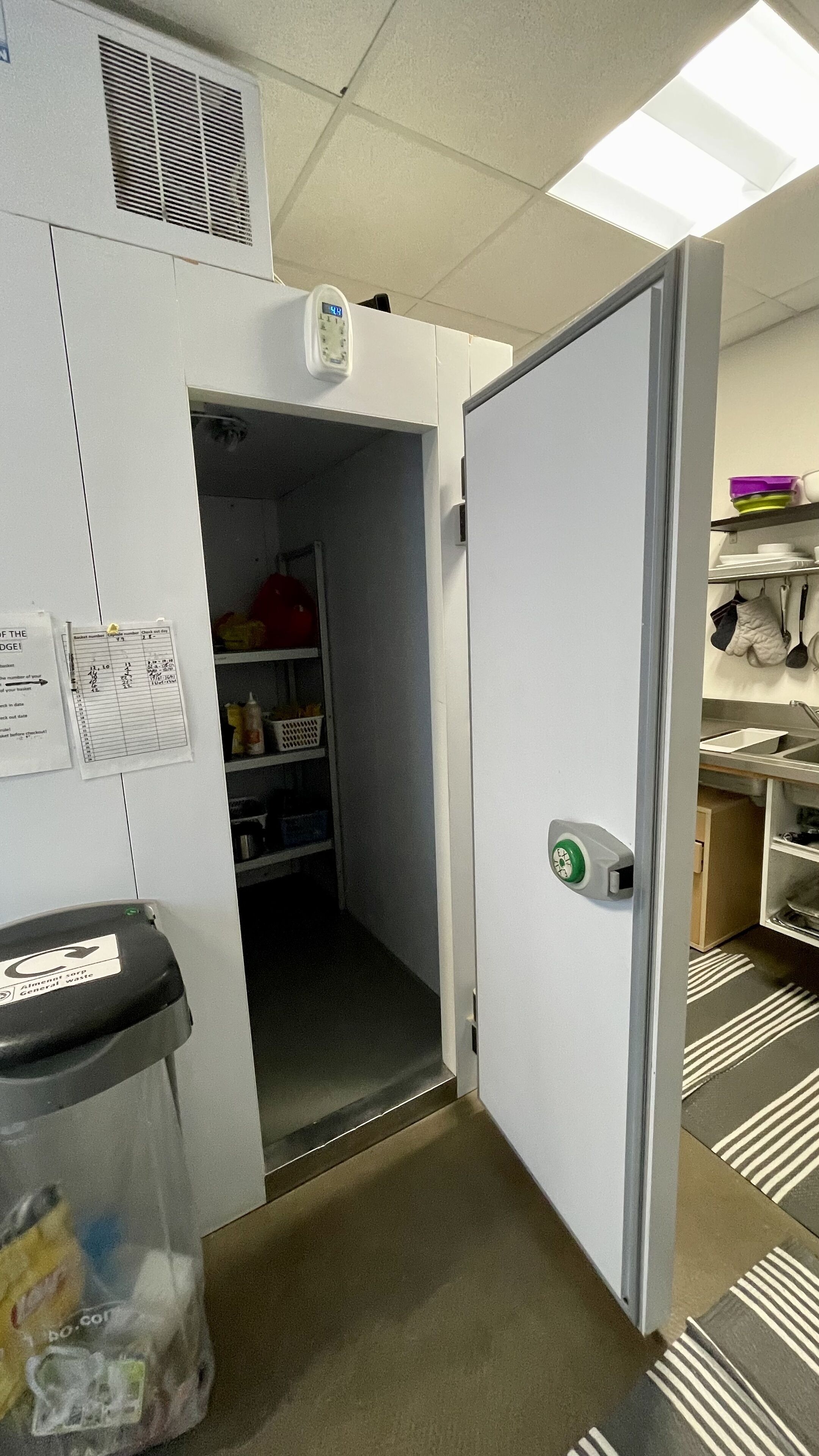 Un pasillo estrecho con un bote de basura, una puerta que da a una zona de cocina y un estante con utensilios de cocina.
