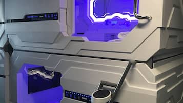 Quarto de hotel moderno em formato de cápsula, com painel de controle central, escada que leva a um nível inferior e interior iluminado em azul.