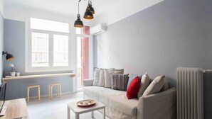 Una sala de estar moderna con un sofá blanco, una mesita pequeña blanca y una ventana con persianas. Hay dos taburetes de madera y un radiador.