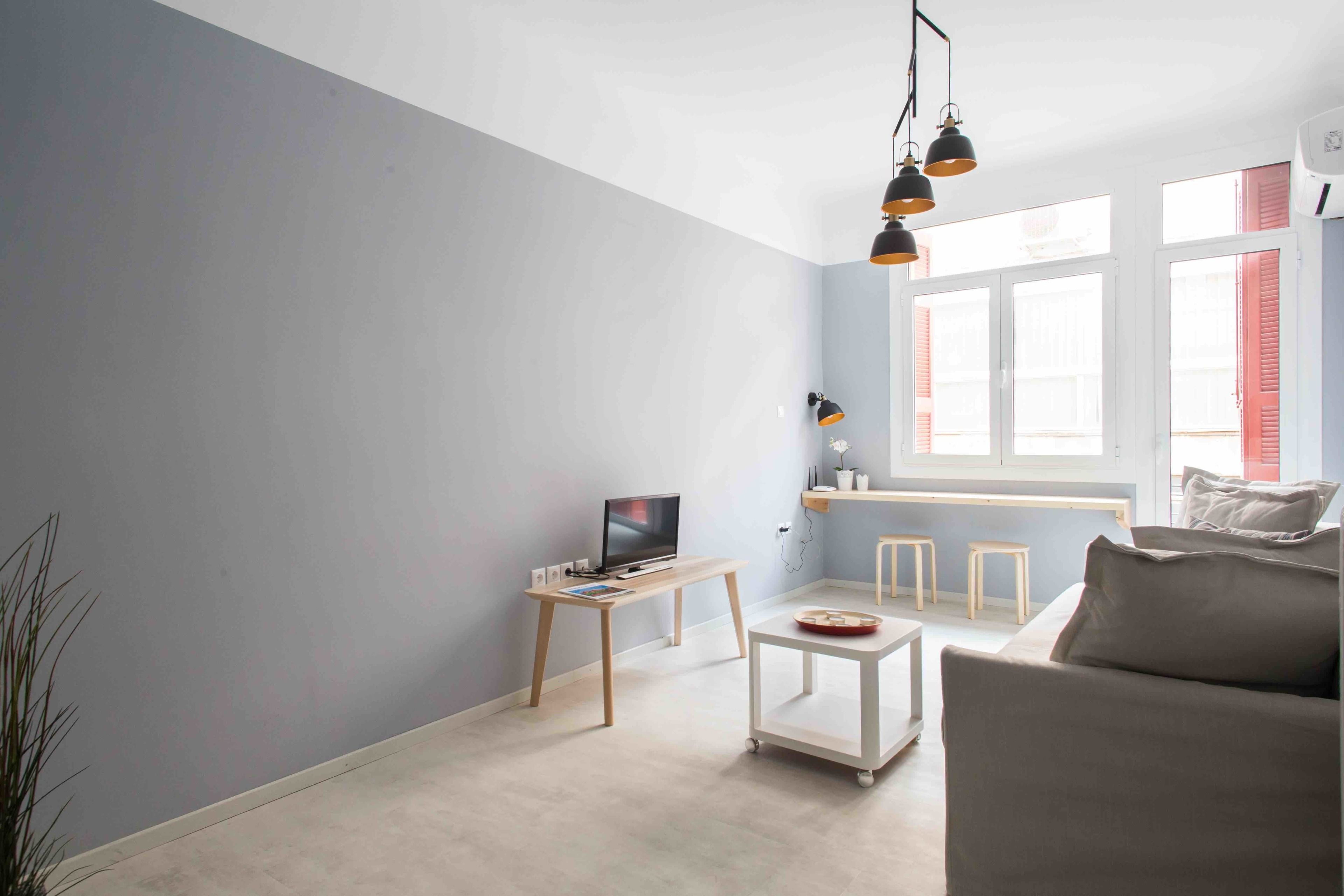 A modern living room with a grey wall, a white sofa, a coffee table, a TV on a wooden stand, and a window with red shutters.