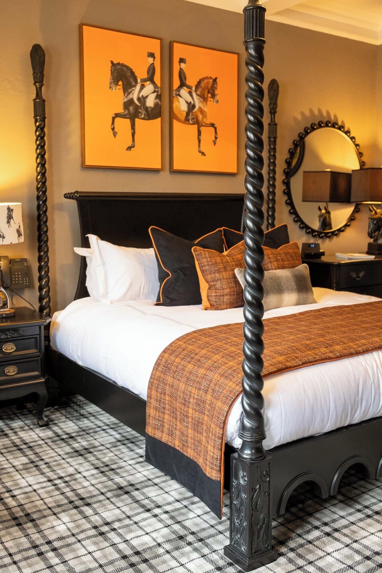 A four-poster bed with a black frame, white bedding, and a brown throw blanket. There are two framed equestrian paintings on the wall above the bed.