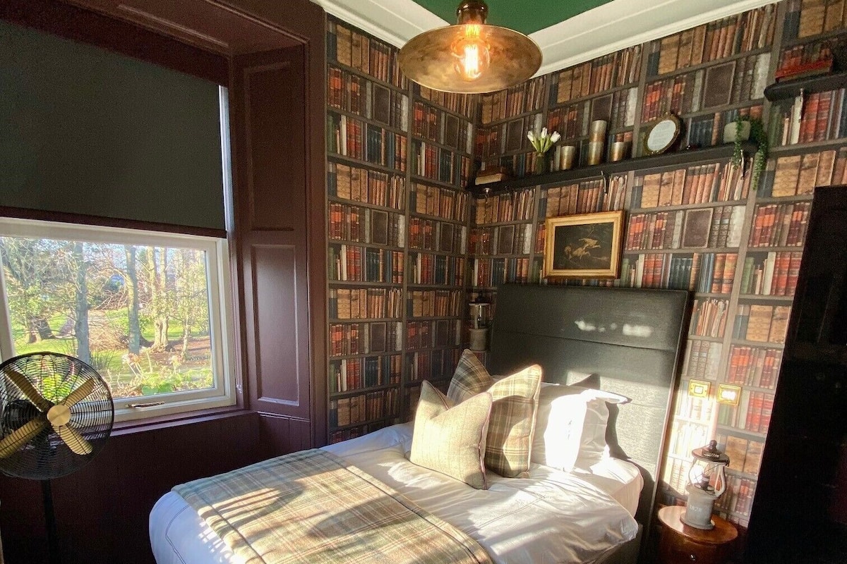 A bedroom with a large bed, a fan, a bookshelf, and a window with a view of trees.