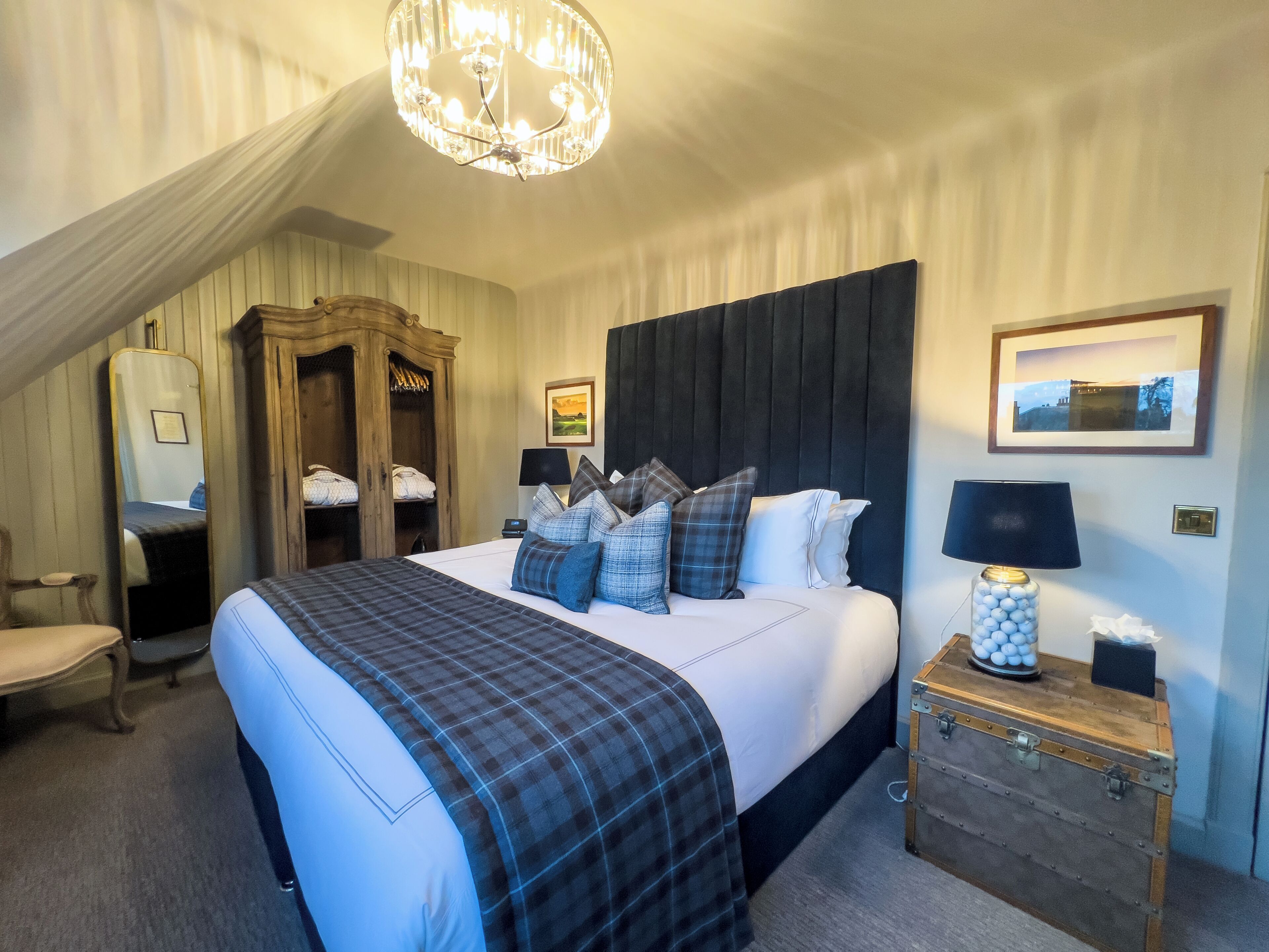 A hotel room with a large bed, a wooden wardrobe, a desk with a lamp, and a chandelier.