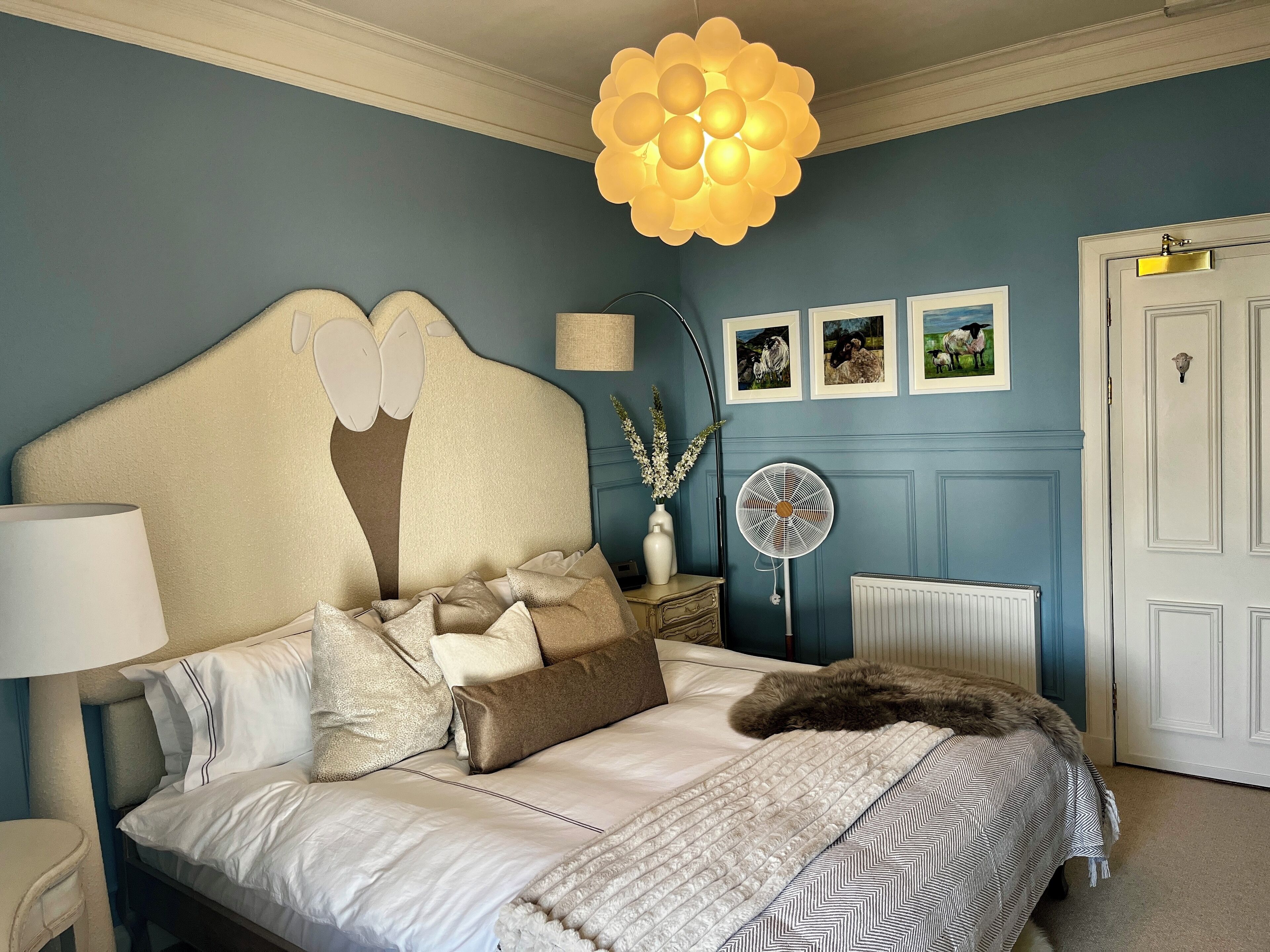 A bedroom with a large bed, a bedside lamp, a floor lamp, a fan, and a door.