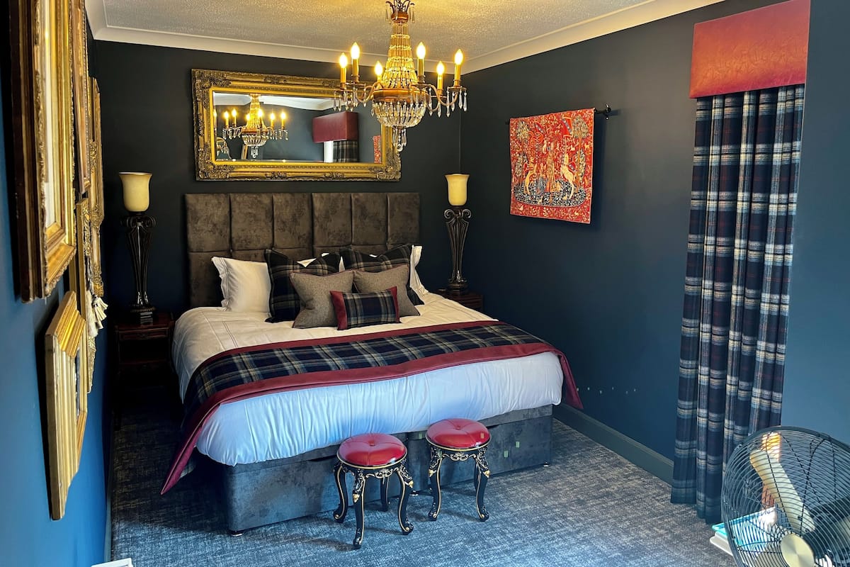 A bedroom with a large bed, a chandelier, a mirror, a red artwork, and a plaid curtain.
