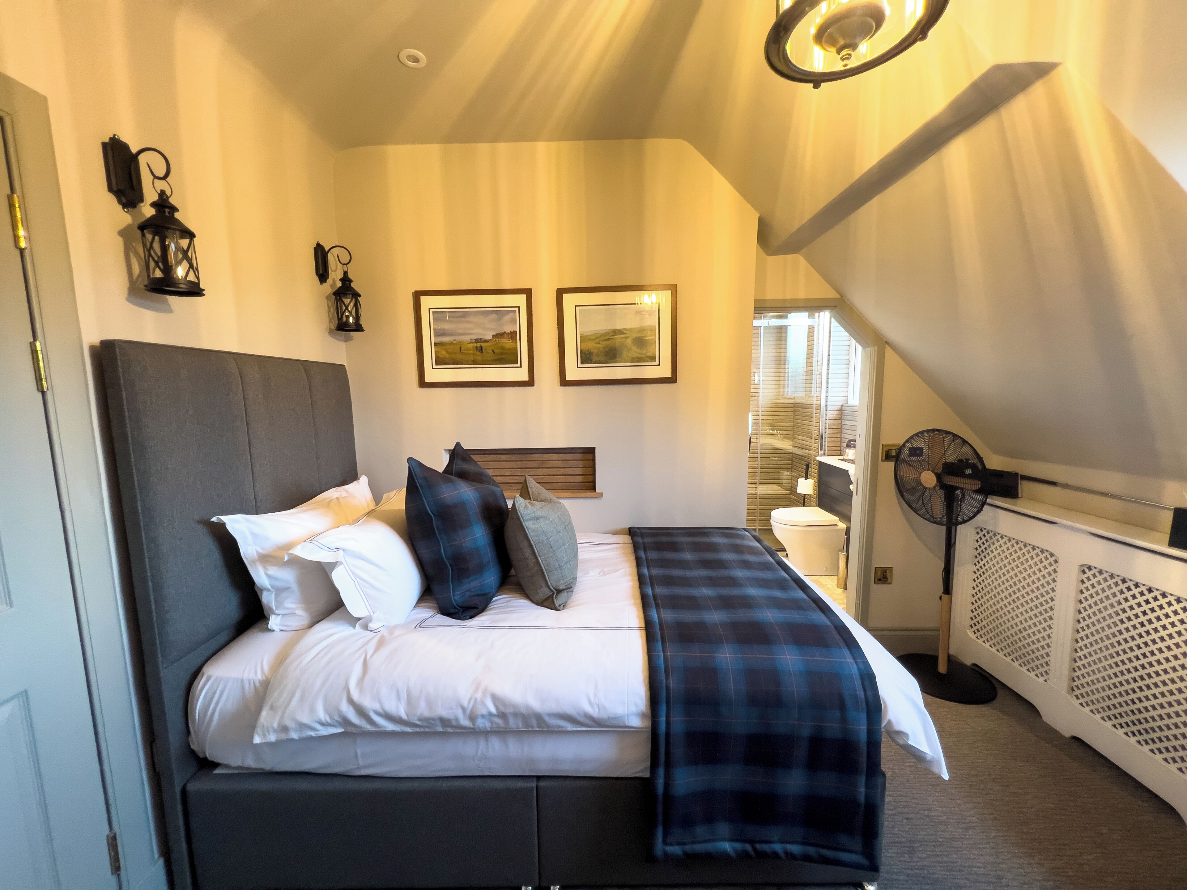 A bedroom with a bed, pillows, a blanket, a standing fan, and two framed pictures on the wall.