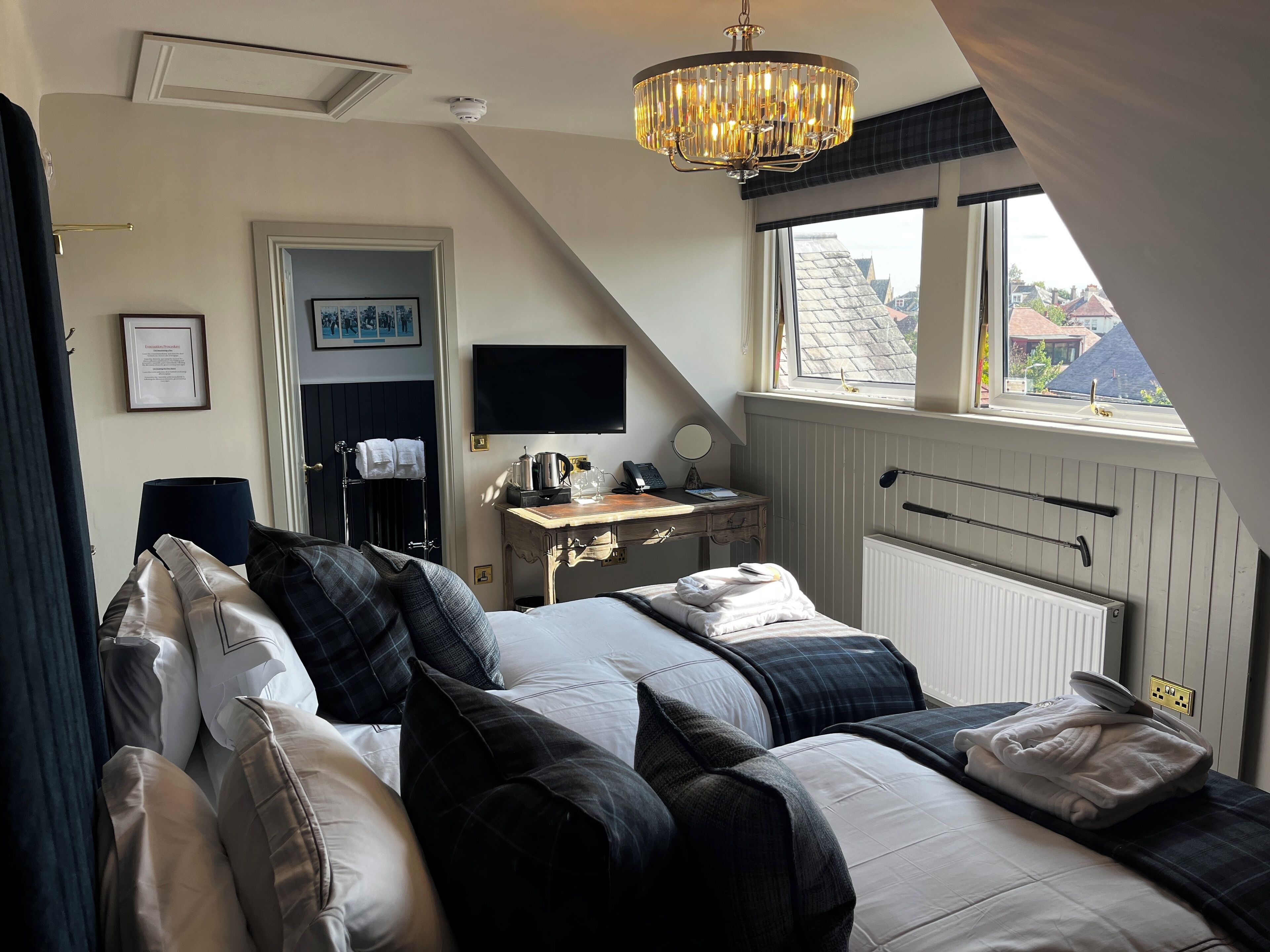 Un dormitorio con cama, escritorio, silla, televisor y ventana con vista a edificios.