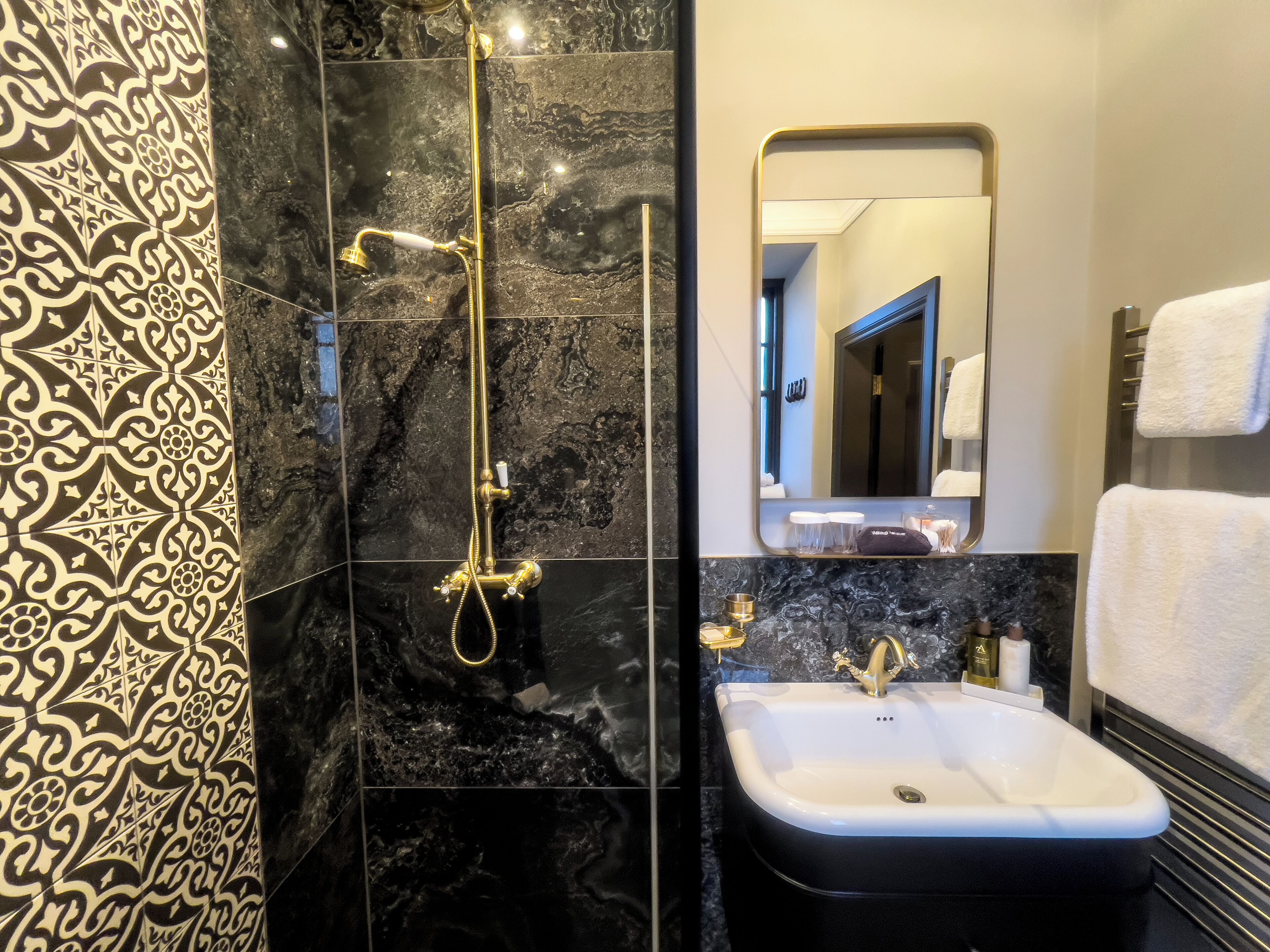 A modern bathroom with a black and gold patterned tile wall, a gold showerhead, and a white sink with a black countertop.