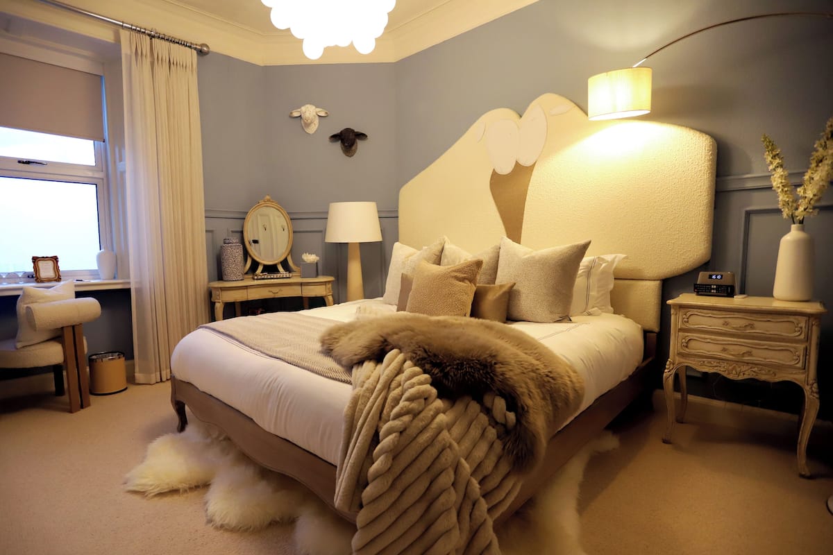 A bedroom with a large bed, a bedside table, a lamp, a mirror, and a window with curtains.