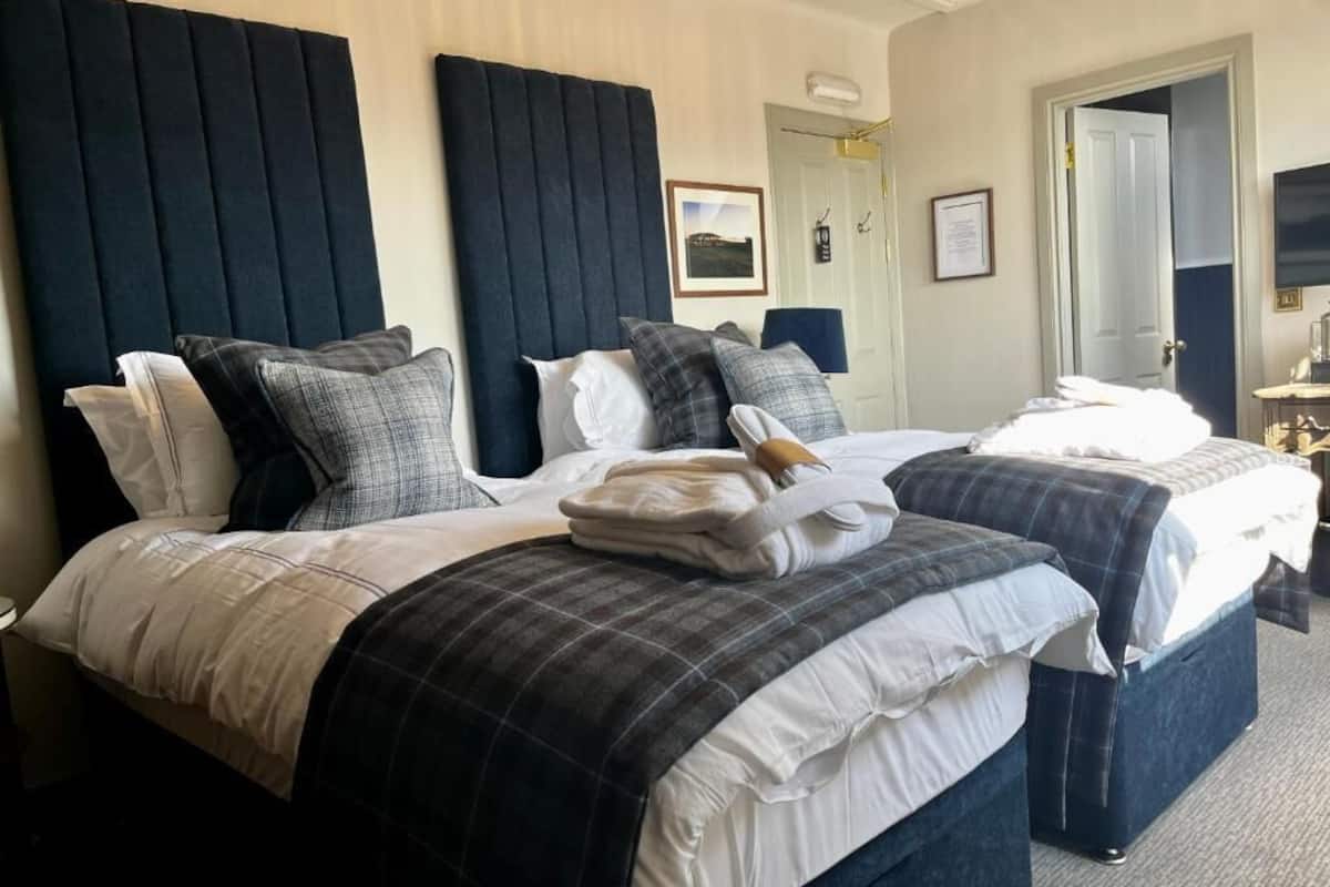 A hotel room with two beds, each with a dark headboard, white bedding, and multiple pillows. There is a door leading to another room and a framed picture on the wall.