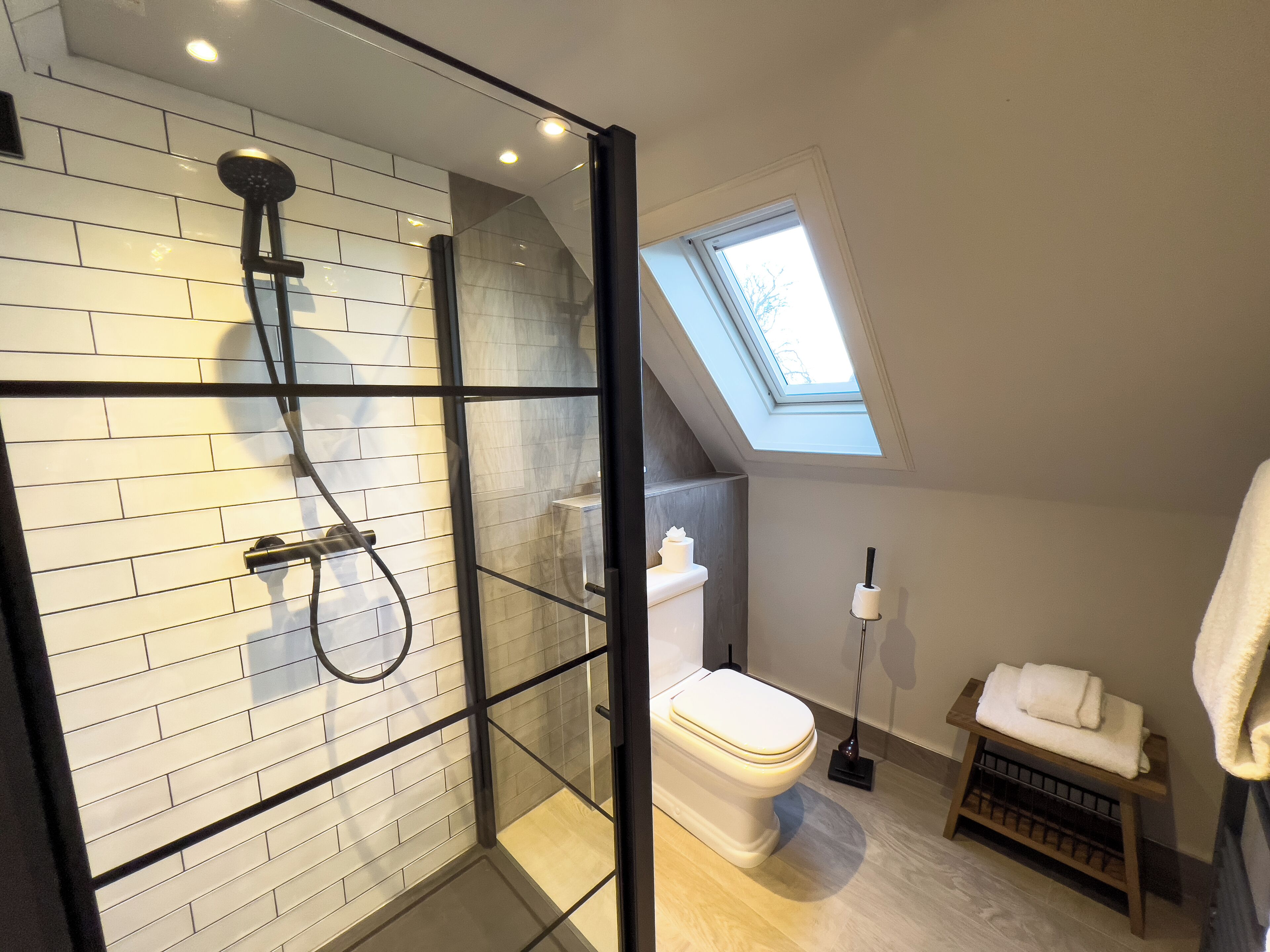 A modern bathroom with a glass-enclosed shower, white tiled walls, a skylight, and a wooden floor.