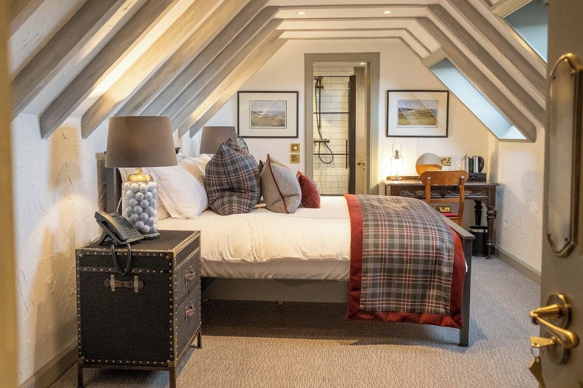 A bedroom with a loft-style ceiling, a bed with a plaid blanket, a desk with a chair, and a suitcase.