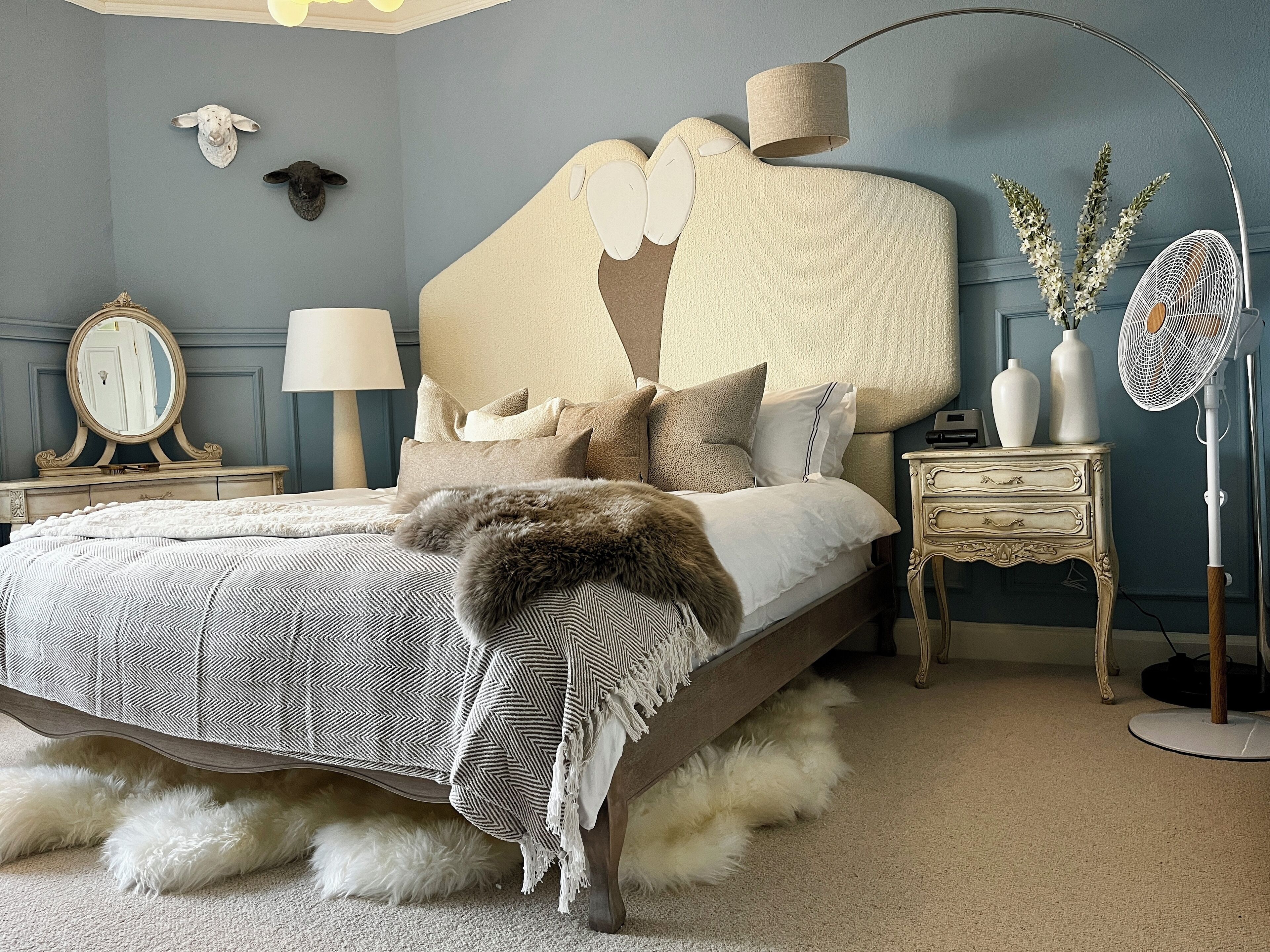 A bedroom with a large bed, a headboard with a sheep design, a lamp, a dresser, and a fan.