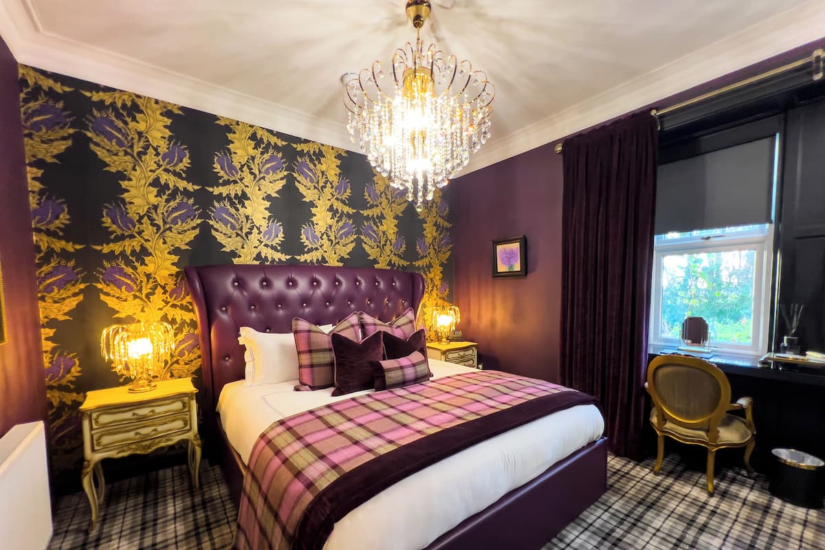 A bedroom with a tufted headboard, a plaid bedspread, a chandelier, a nightstand with a lamp, a chair, and a window with curtains.