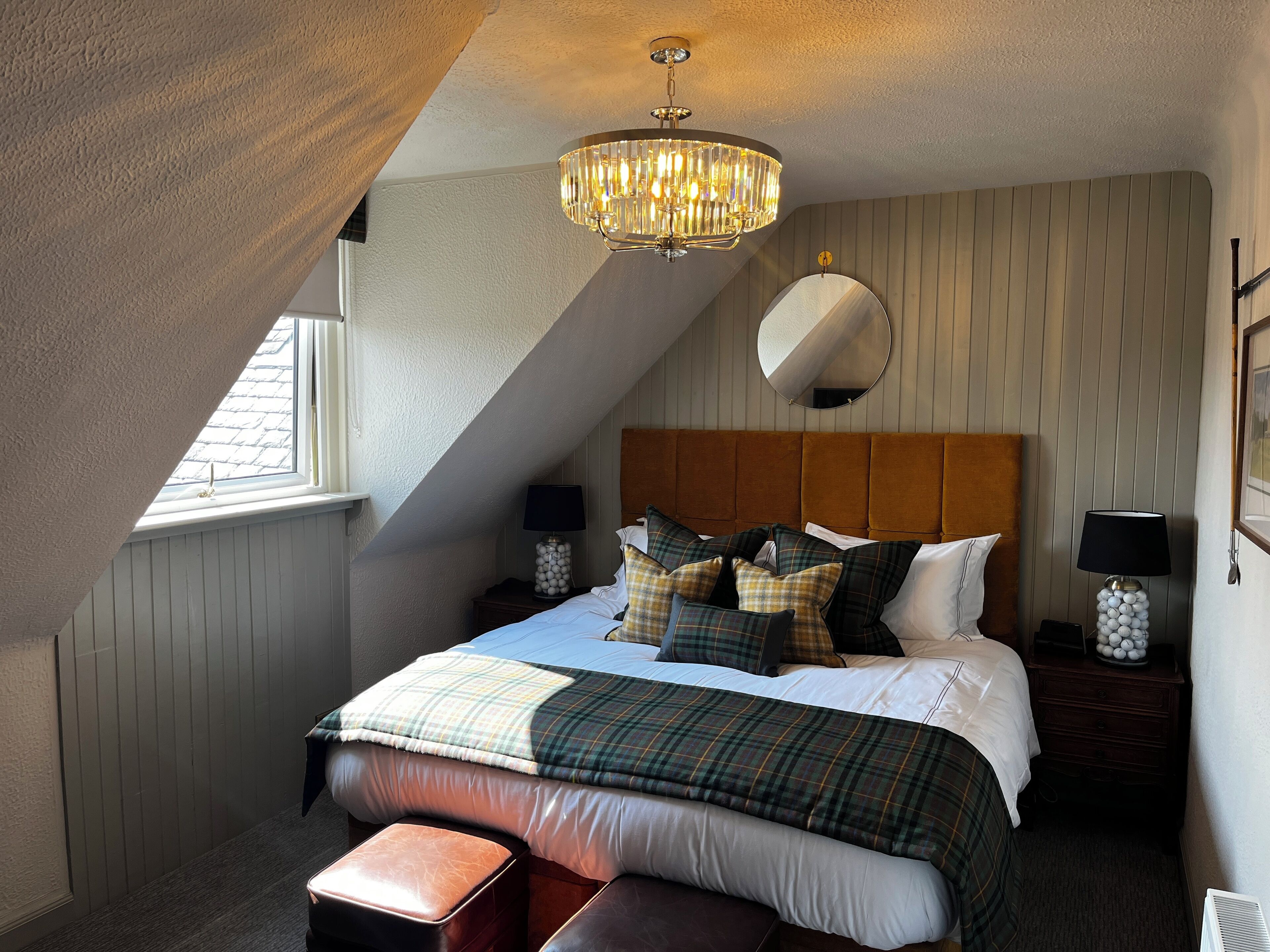 Ein Schlafzimmer mit schrägen Wänden, einem großen Bett mit vielen Kissen, einem Nachttisch mit Lampe, einem Spiegel und einem Fenster.