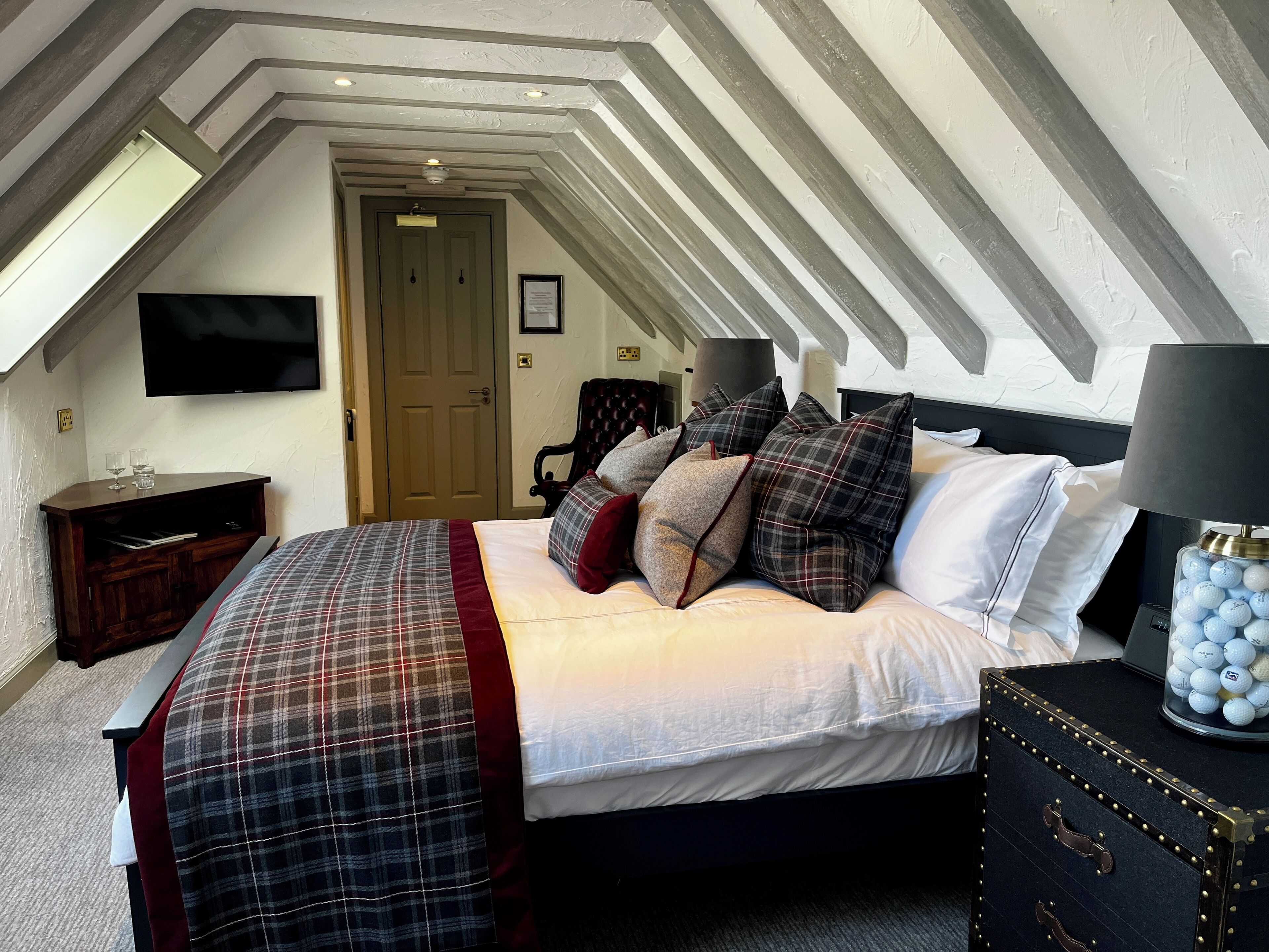 A bedroom with a sloped ceiling, a bed with plaid bedding, a nightstand, a lamp, and a decorative container.