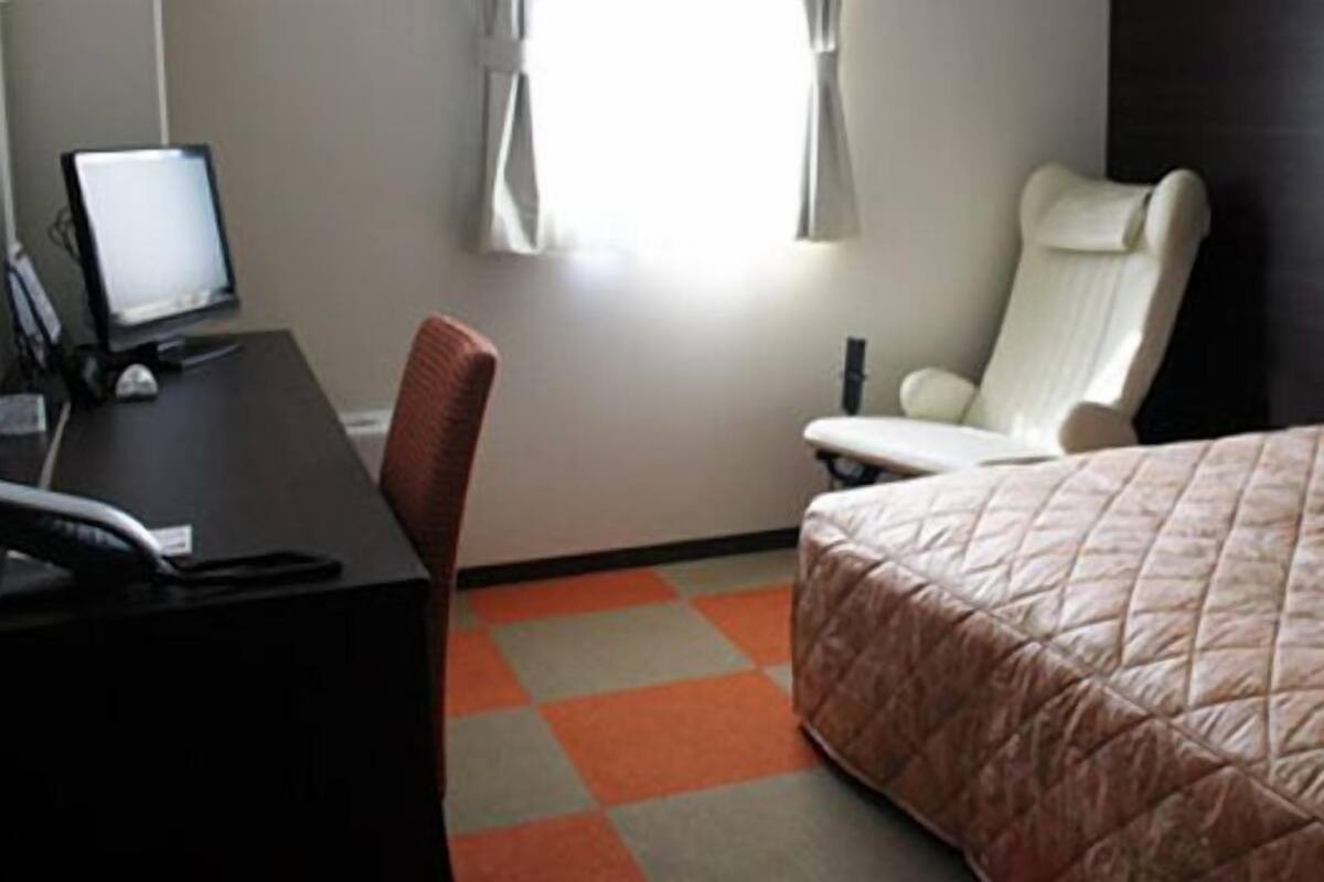 Ein Hotelzimmer mit einem Bett, einem Schreibtisch mit Computer, einem Sessel und einem Fenster mit Vorhängen.