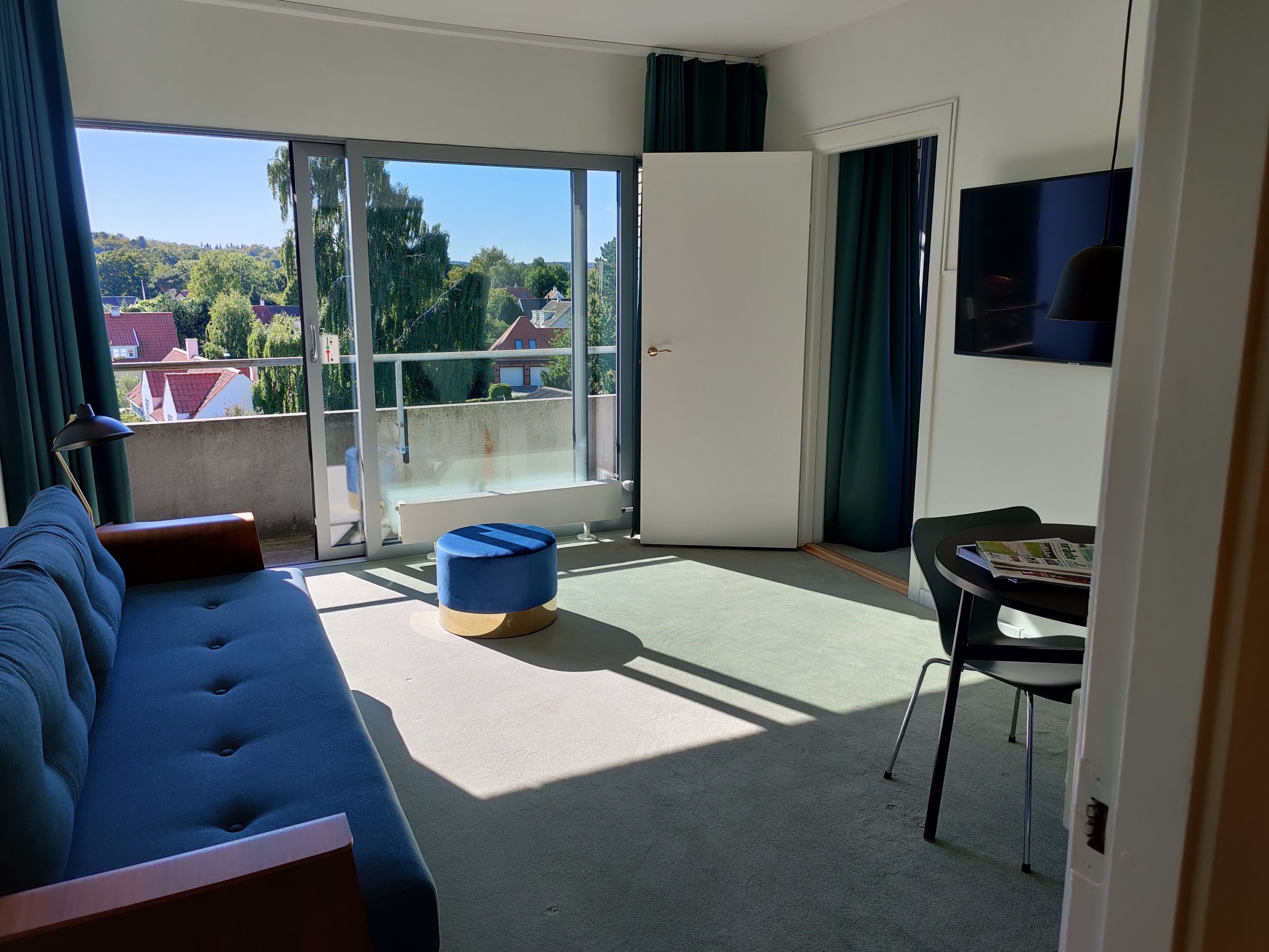 Una sala moderna con un sofá, una mesita y una silla. Hay un ventanal corredero que da a un balcón con vistas a casas y árboles.
