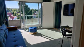 Una sala moderna con un sofá, una mesita y una silla. Hay un ventanal corredero que da a un balcón con vistas a casas y árboles.