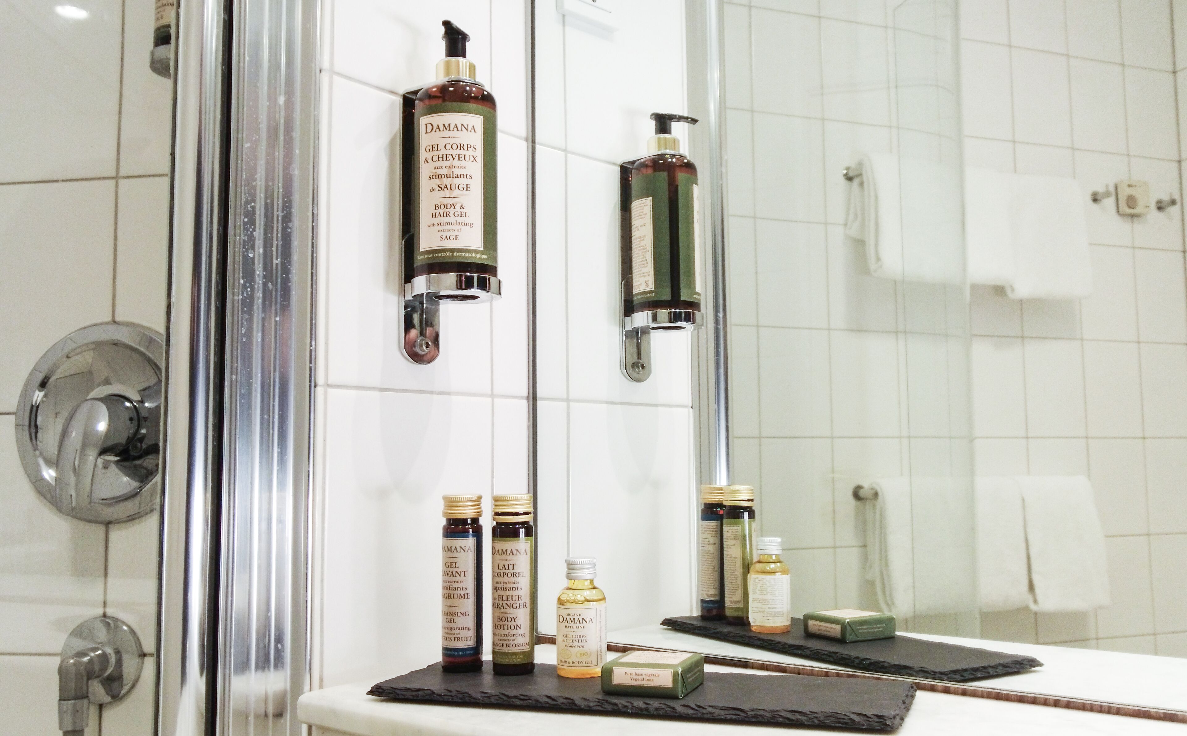 A bathroom with a glass shower door, white tiled walls, and a mirror reflecting the room. On the wall, there are several bottles of Damana body care products, including gel, lotion, and body lotion.